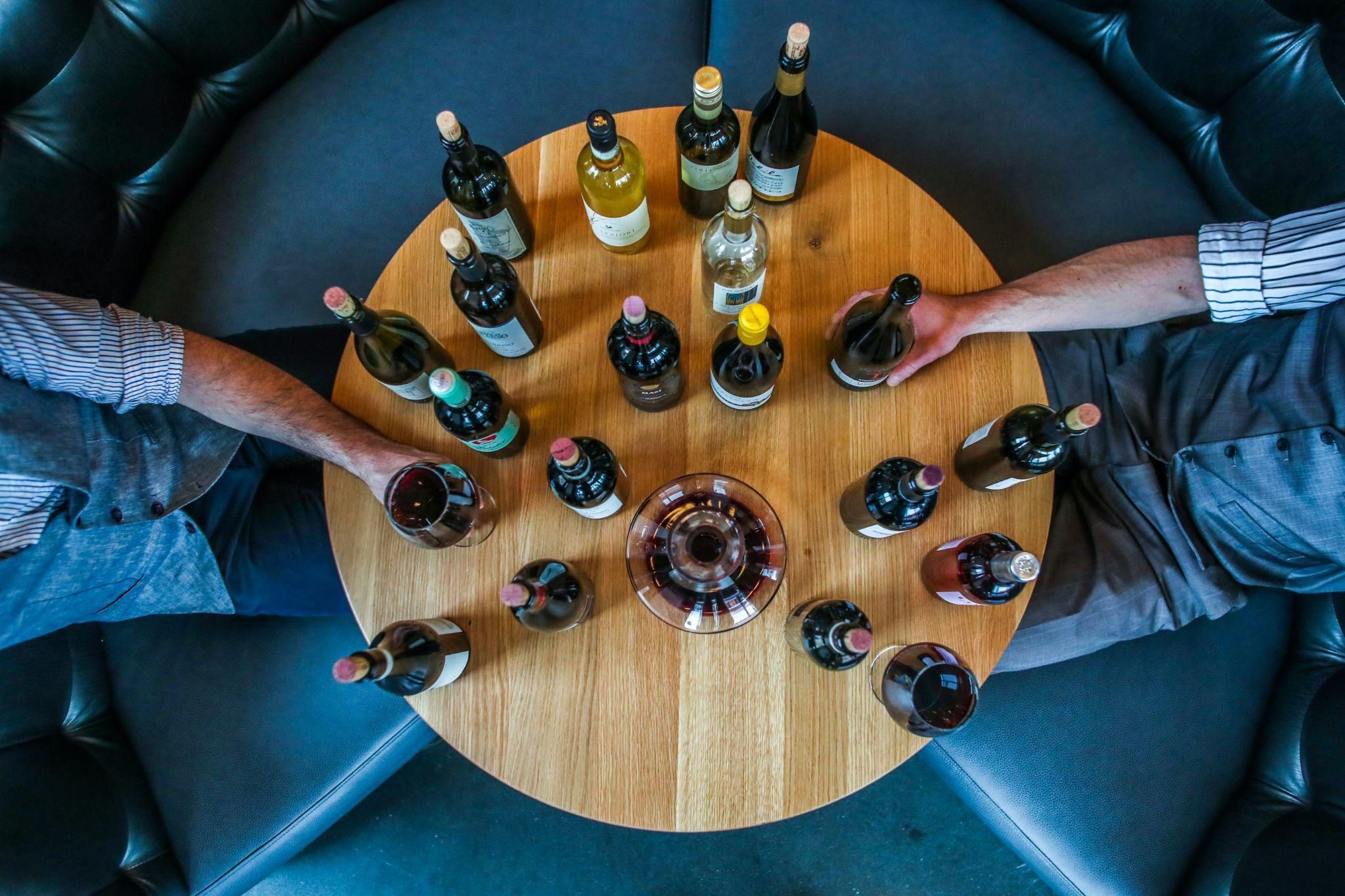 Two men sitting face to face at a circular table on which there are over a dozen bottles of wine