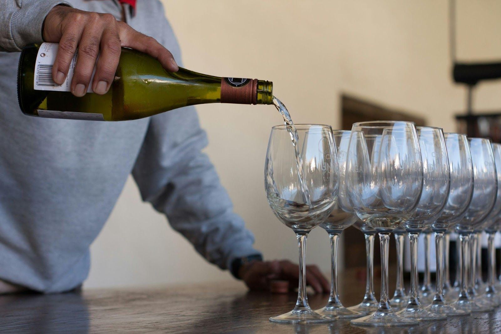 A person pouring white wine into a row of wine glasses on a counter